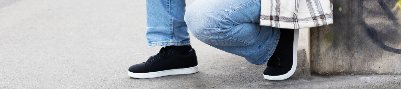 Person in Jeans und kariertem Hemd hockt an einer Wand und trägt schwarze Turnschuhe auf einem gepflasterten Untergrund.