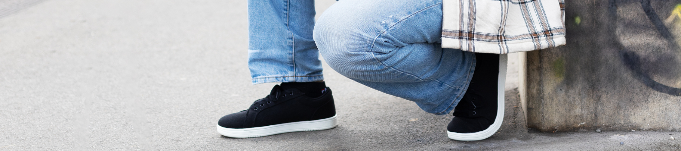 Person in Jeans und schwarzen Turnschuhen kauert auf dem Bürgersteig neben einer Graffitiwand.