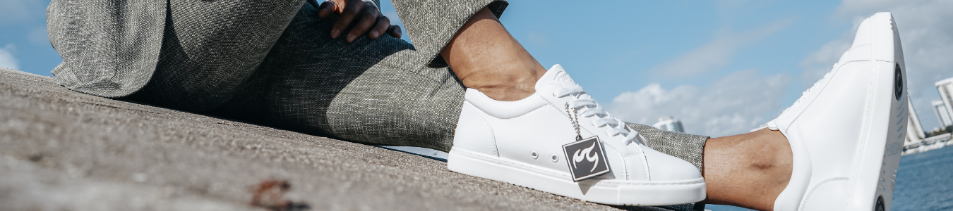 Person in grauer Hose und weißen Turnschuhen, die auf einer geneigten Betonfläche vor dem Hintergrund eines Himmels und einer Stadtlandschaft sitzt.