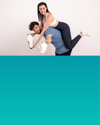 Ein Mann hält eine Frau auf seiner Schulter, beide lächeln, während sie mit einem Turnschuh in die Kamera zeigt.