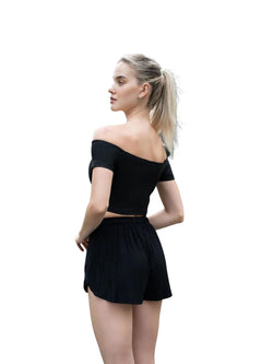 Person in Lisminas 084 Comfortlux Off Shoulder T-Shirt in Schwarz und Shorts, die vor einem weißen Hintergrund posiert.