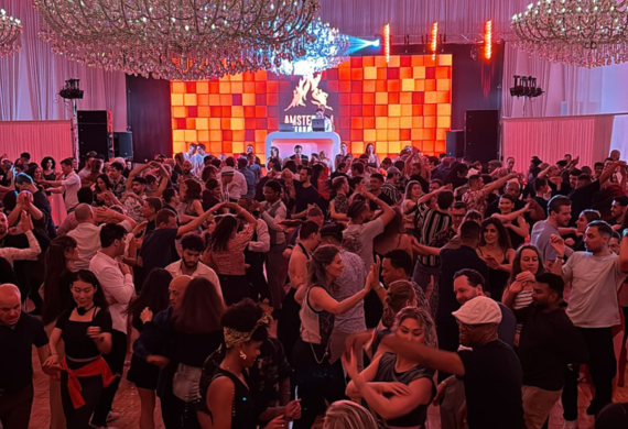 Eine große Menschenmenge tanzt energisch in einem Veranstaltungsort mit Kronleuchtern und einem farbenfrohen DJ-Pult im Hintergrund.