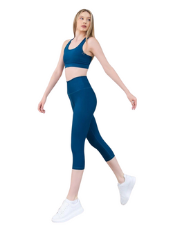 Person in Lisminas petrolblauen 4314 Capri-Leggings und weißen Sneakers, die selbstbewusst vor einem weißen Hintergrund posiert.