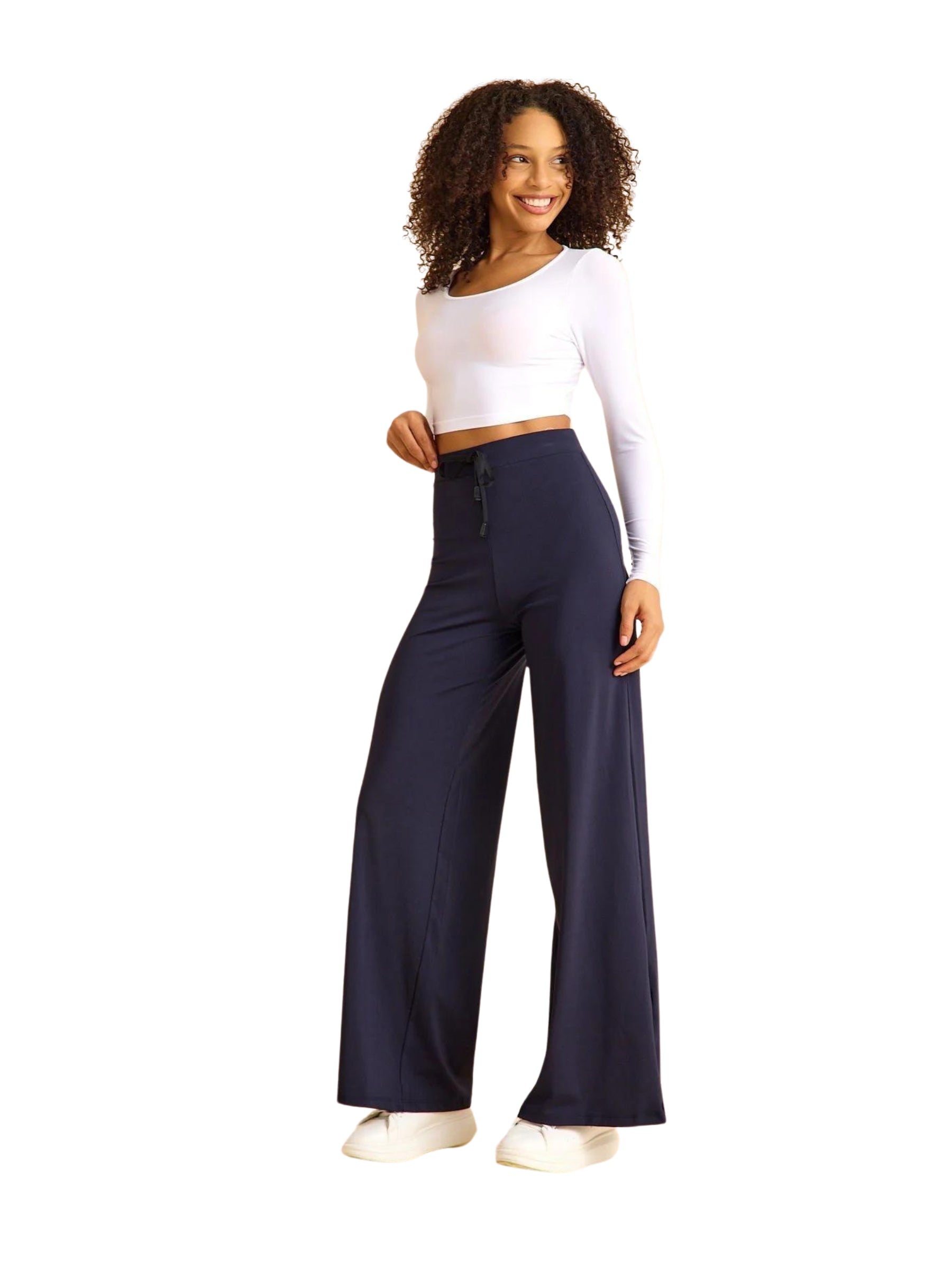 Person in einem weißen bauchfreien Top, der marineblauen 447 Mumbai Yoga Hose von Lismina und weißen Turnschuhen, lächelt vor einem schlichten Hintergrund.
