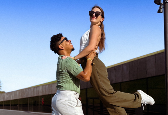 Ein Mann hebt im Freien unter einem klaren blauen Himmel spielerisch eine lächelnde Frau hoch.