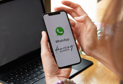 Hände halten ein Telefon mit dem WhatsApp-Logo und dem Text „Dancing Queens“, im Hintergrund ist ein geöffneter Laptop zu sehen.