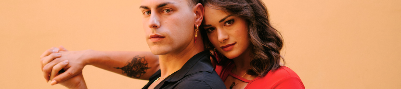 Ein ernst dreinblickendes Paar posiert eng beieinander vor einem schlichten pfirsichfarbenen Hintergrund und hält sich an den Händen.