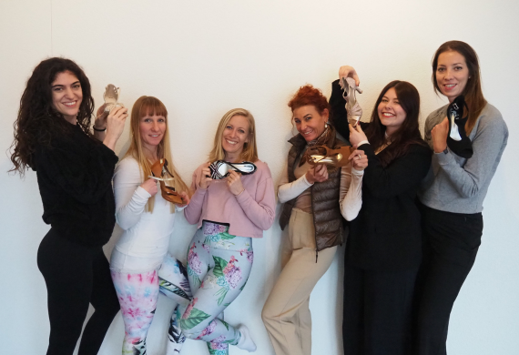 Sechs Frauen posieren fröhlich vor einer weißen Wand und halten schicke Stöckelschuhe in den Händen.