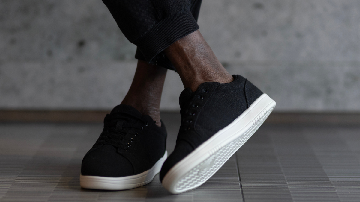 Person, die schwarze Turnschuhe mit weißen Sohlen trägt und auf einem gefliesten Boden steht.