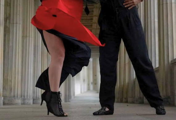 Zwei Personen tanzen, die eine in einem roten Kleid und hochhackigen Schuhen, die andere in schwarzen Hosen und Schuhen, in einem Säulengang.