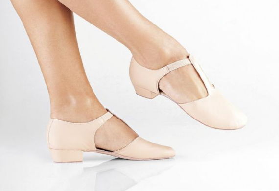 Beine in hellrosa Tanzschuhen mit niedrigem Absatz und T-Riemen, abgebildet vor weißem Hintergrund.