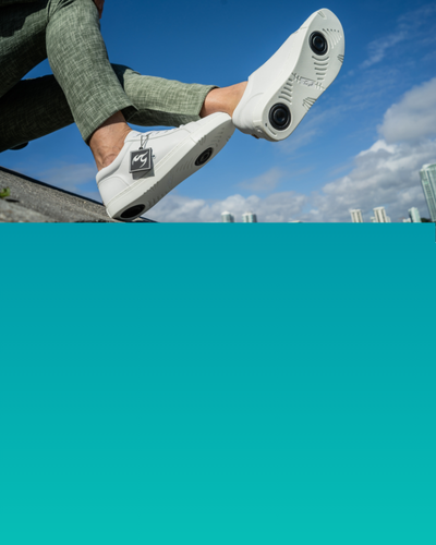 Person in weißen Turnschuhen mit kreisförmigen Mustern auf den Sohlen, die im Freien vor einem blauen Himmel sitzt.