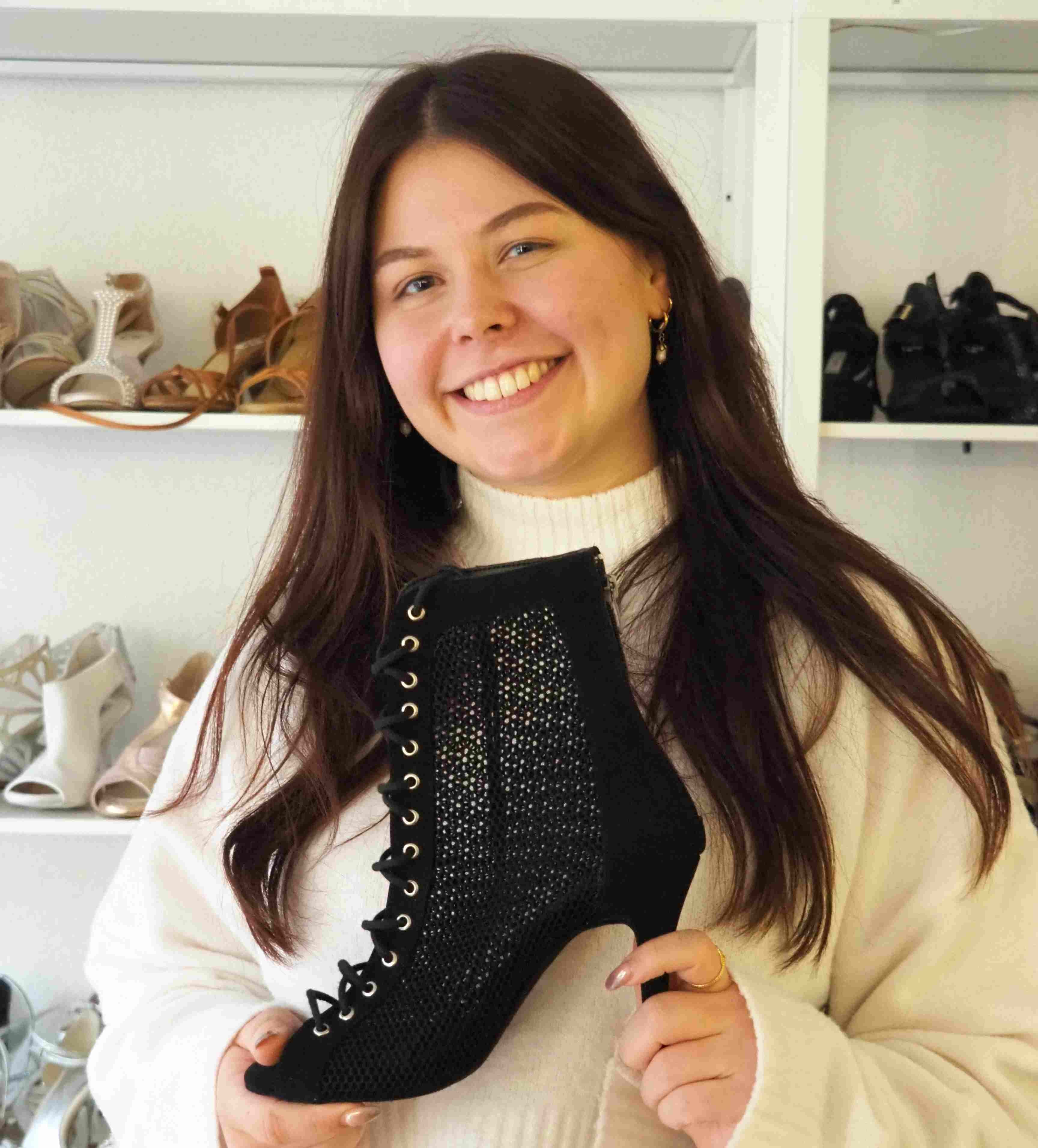 Eine lächelnde Frau hält einen schwarzen Schnürschuh in einem Geschäft, hinter ihr stehen Regale mit verschiedenen Schuhen.