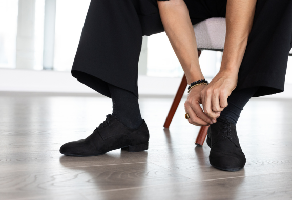 Person, die auf einem hellen Holzboden sitzt und schwarze Schuhe bindet, schwarze Socken und Hosen trägt.