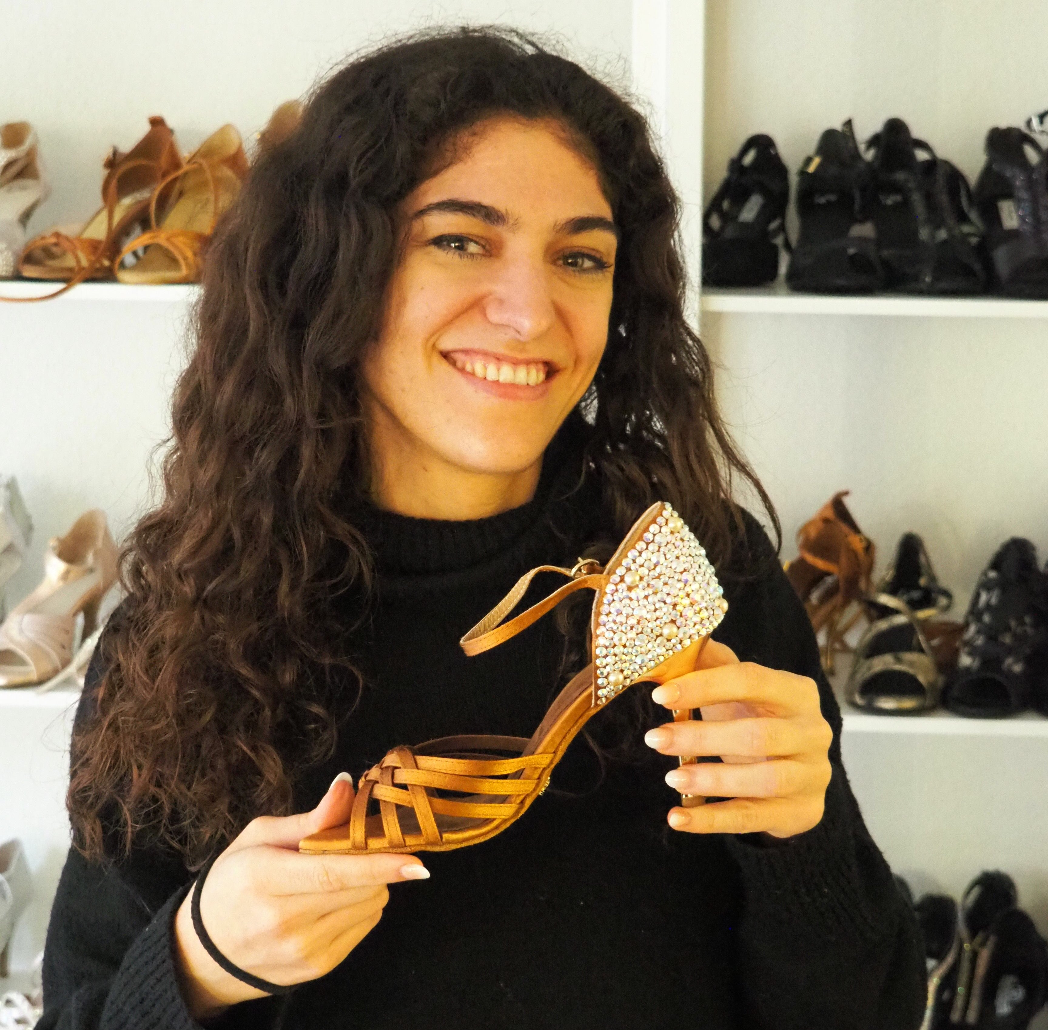 Eine Person lächelt und hält eine Riemchensandale vor einem Schuhregal.