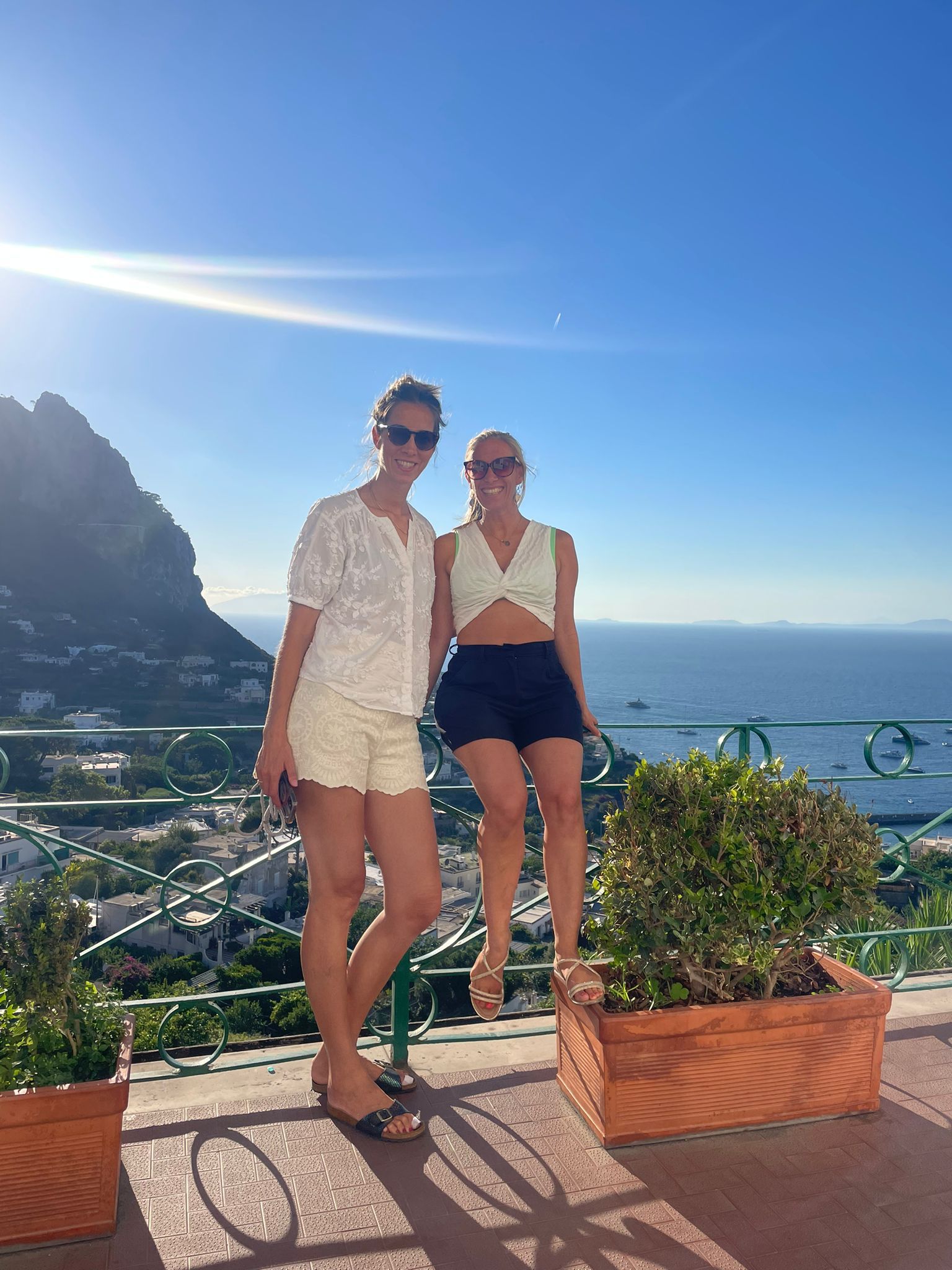 Zwei lächelnde Frauen auf einer sonnigen Terrasse mit Blick auf das Meer und die Berge im Hintergrund.