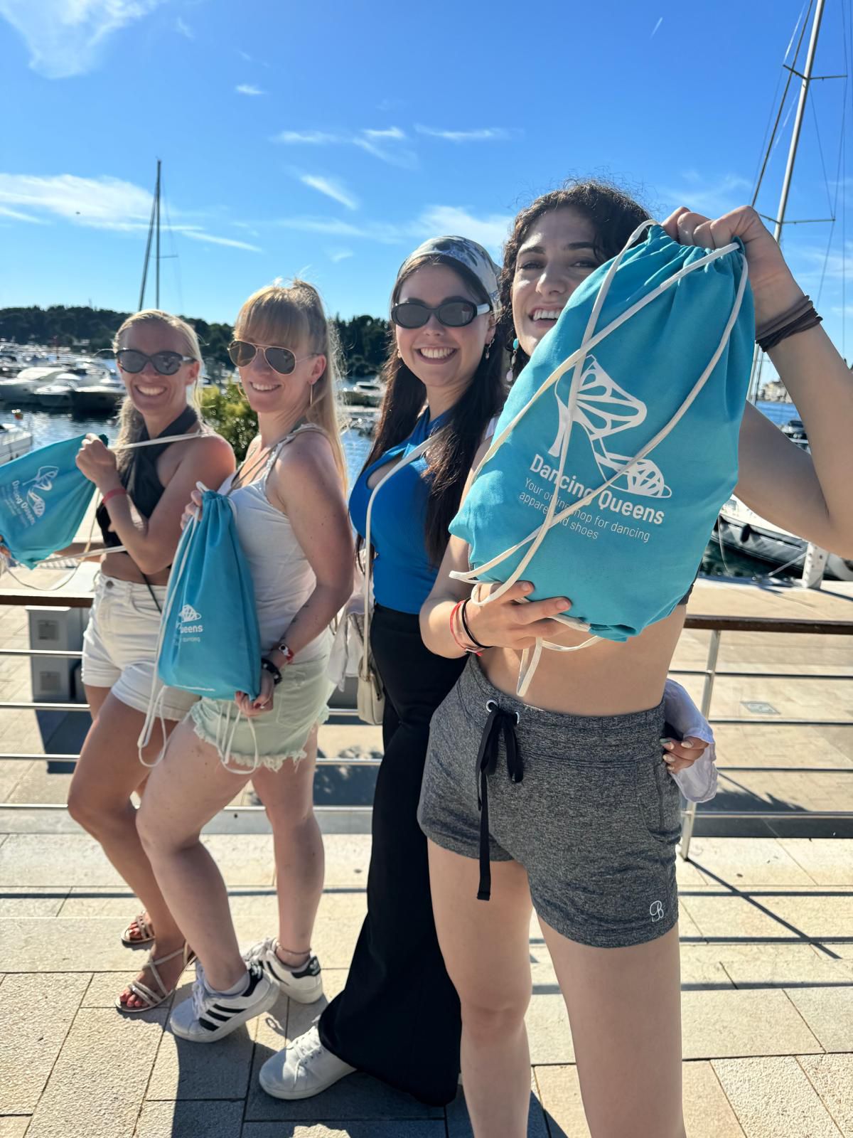 Vier lächelnde Menschen halten blaue Beutel mit Kordelzug in der Hand und stehen auf einem Pier mit einem Jachthafen im Hintergrund.