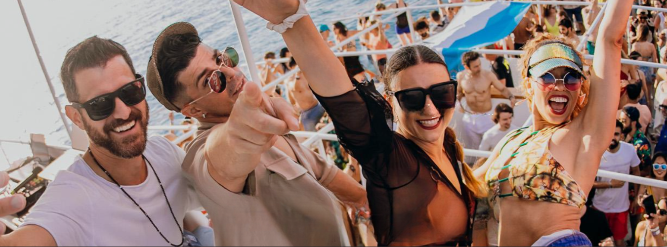 Vier Freunde mit Sonnenbrillen lächeln und tanzen auf einer lebhaften Strand- oder Bootsparty im Freien mit einer Menschenmenge hinter ihnen.