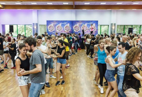 Eine große Gruppe von Menschen tanzt paarweise in einem hellen Hallenstudio mit Holzfußboden und bunten Bannern.