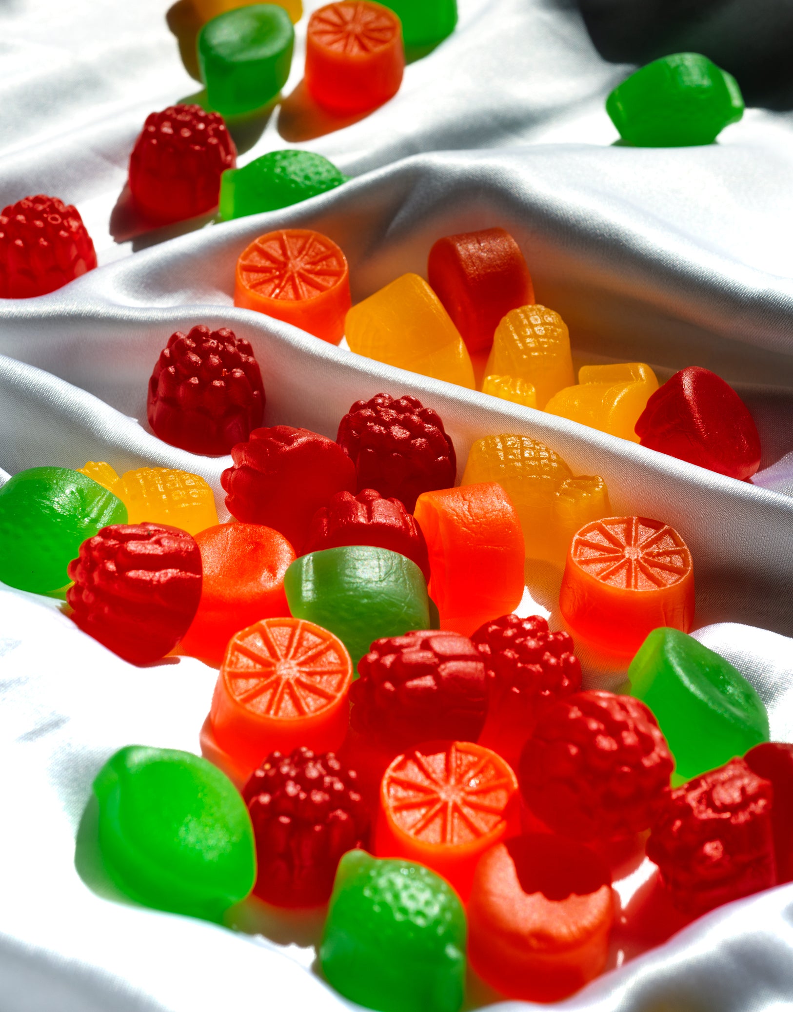 Bunte, fruchtförmige Gummibonbons auf weißem, seidenem Stoff in hellem Sonnenlicht verstreut.