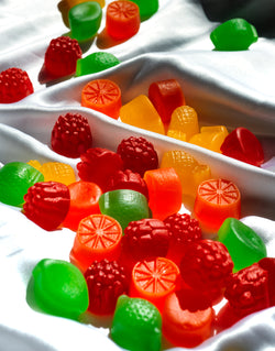 Bunte, fruchtförmige Gummibonbons auf weißem, seidenem Stoff in hellem Sonnenlicht verstreut.