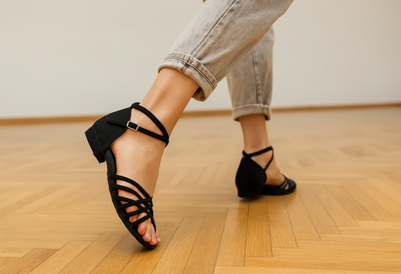 Eine Person in schwarzen Riemchensandalen und hochgekrempelten Jeans läuft auf einem hellen Holzboden.