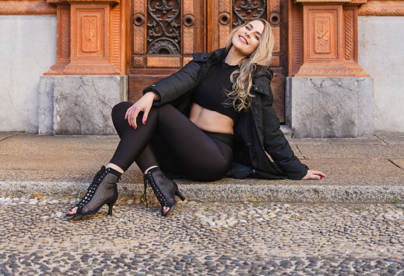 Eine Frau in schwarzem Outfit sitzt draußen auf dem Boden und lächelt, hinter ihr befindet sich eine verzierte Tür.