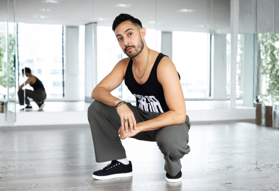 Ein Mann in einem schwarzen Tanktop und einer grauen Hose hockt in einem hellen, modernen Tanzstudio mit verspiegelten Wänden.
