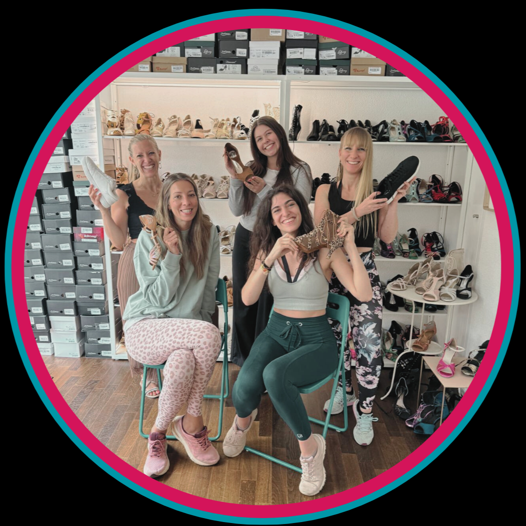 Fünf lächelnde Frauen mit Schuhen in der Hand sitzend und stehend in einem Raum mit Regalen voller Schuhkartons.