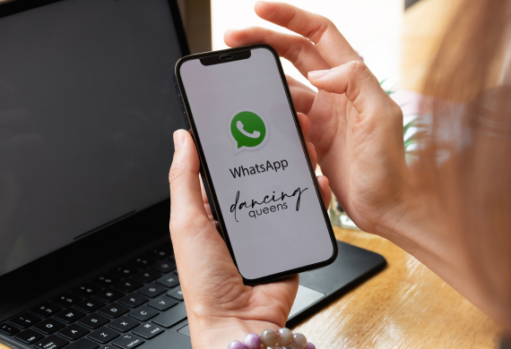 Hände halten ein Telefon mit dem WhatsApp-Logo und dem Text „Dancing Queens“, im Hintergrund ist ein geöffneter Laptop zu sehen.