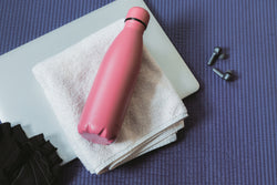 Eine rosa Wasserflasche auf einem weißen Handtuch, mit Kopfhörern und einer Yogamatte im Hintergrund.