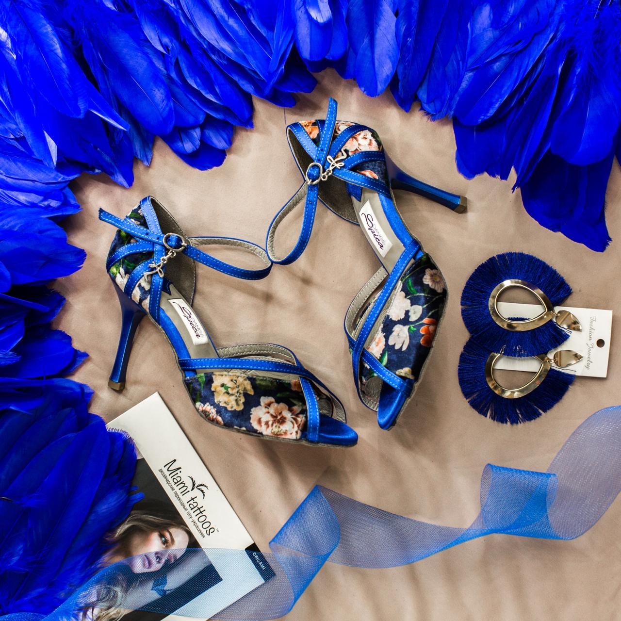 Naos Tanzschuhe in Blau geblumt von Dancing Queens mit passenden Ohrringen auf Beige, umgeben von blauen Federn.