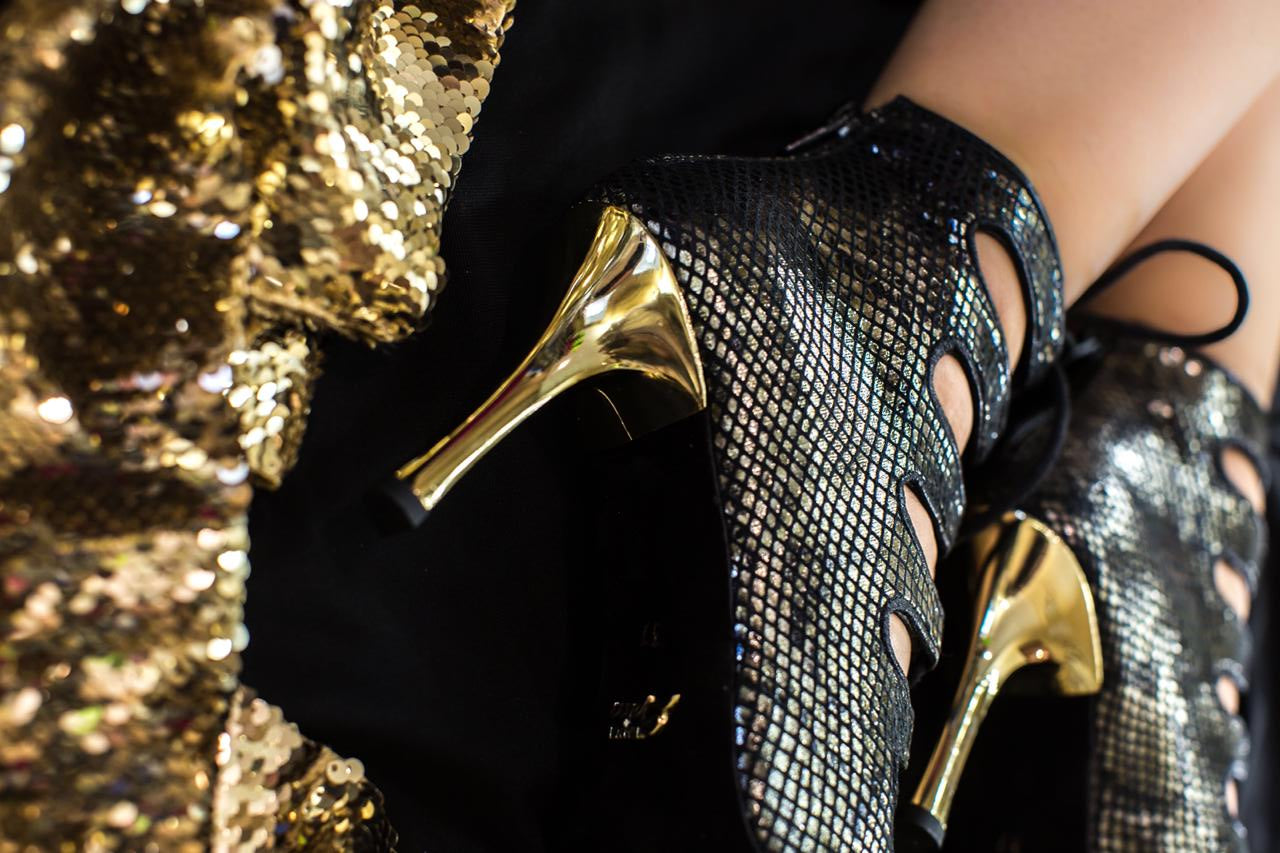Nahaufnahme der schwarzen Piton-Electra-Tanzschuhe von Dancing Queens mit goldenen Absätzen neben goldenem Paillettenstoff.